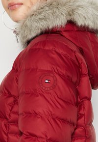 Close-up van een rode pufferjack met een met bont afgewerkte capuchon en een ronde Tommy Hilfiger-logo patch op de bovenste mouw.