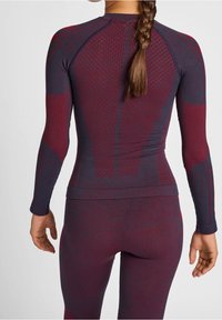 Nainen, jolla on letitetyt hiukset, pukeutuneena istuvaan pitkähihaiseen puseroon ja leggingseihin, joissa on tekstuuriset laivastonsiniset ja viininpunaiset kuviot, kuvattuna takaa.