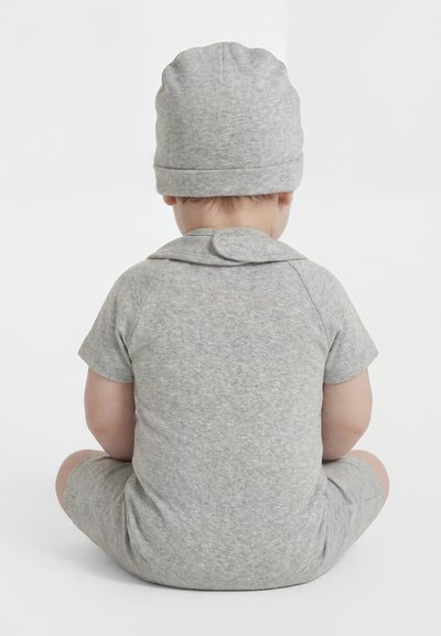 Niemowlę ubrane w szary kombinezon z krótkim rękawem i dopasowaną czapkę, siedzące ze skrzyżowanymi nogami tyłem na jednolitym białym tle.