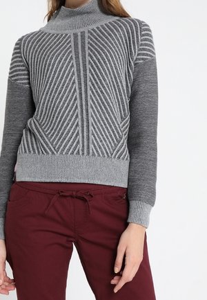 Femme portant un pull à col roulé gris avec des rayures diagonales texturées et des manches unies, associé à un pantalon bordeaux à cordon de serrage.