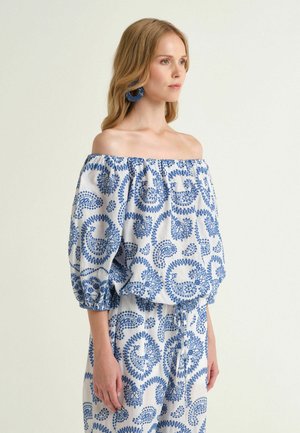 Femme portant un top blanc épaules dénudées et un pantalon avec des motifs cachemire bleus et des boucles d’oreilles rondes bleues assorties, debout devant un fond uni.