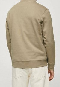 Sweat-shirt beige à encolure ronde avec poignets et ourlet côtelés. Texture lisse, design minimaliste, présenté de dos avec un pantalon de couleur claire.