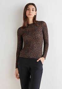 Vrouw in een nauwsluitend, lang mouwend leopardprint shirt en zwarte broek, die met één hand in haar zak tegen een effen lichte muur staat.