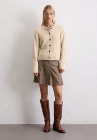 Kremowy kardigan o teksturze z guzikami, zestawiony z brązową plisowaną mini spódnicą w kratę i brązowymi skórzanymi kozakami do kolan.