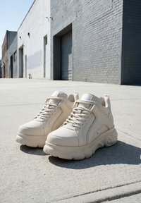Kraftige beige sneakers med tyk sål, placeret på en solbeskinnet urbane fortov foran en grå murstensbygning med lukkede garagedøre.