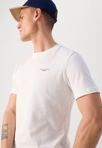 Witte katoenen t-shirt met korte mouwen en een ronde halslijn. Heeft een klein zwart logo op de borst. Glad textuur.