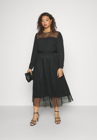 Svart kjole med lange ermer, gjennomsiktig spissbaldyr og en plisserte tylskjørt. Kombinert med svarte stroppehæler og en liten svart clutch.