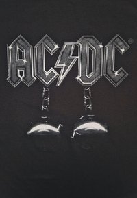 Černé tričko s velkým, výrazným logem "AC/DC" v kovově stříbrných akcentách a dvěma stříbrnými zvonky visícími pod ním. Obsahuje texturované designové prvky.