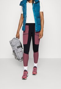 Vadderad teal väst, vit t-shirt, korg med vinröda och svarta leggings med dragkedjor, grå ryggsäck, rosa vandringsskor med vita sulor.