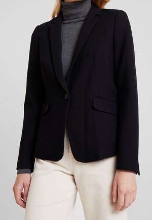 Vrouw draagt een zwart eenknopsblazer over een grijze coltrui en crèmekleurige broek, staand tegen een effen achtergrond.