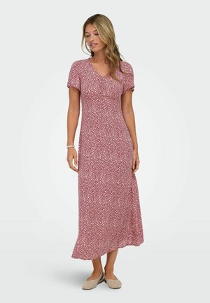 Kvinde iført rød og hvid blomstrede maxi-kjole med korte ærmer og beige flettede flade sko, stående mod en ensfarvet lys baggrund.