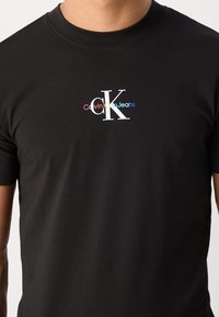 Svart bomullströja med en flerfärgad "Calvin Klein Jeans"-logotyp och stiliserad "CK" på bröstet, rund halsringning och korta ärmar.