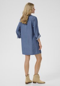 Jeanshemdkleid mit langen Ärmeln, umgeschlagenen Manschetten und legerem Schnitt. Hellblaue Farbe und roher Saum. Getragen mit beigen Stiefeletten.