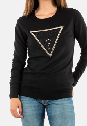 Femme portant un t-shirt à manches longues noir avec un triangle du logo Guess orné de strass et un jean bleu, debout avec une main dans la poche.