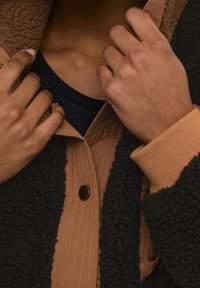 Musta fleecepusero, jossa on ruskea tikattu kaulus, resorit hihansuissa ja kultaiset painonapit. Musta aluskerros näkyvissä alla.