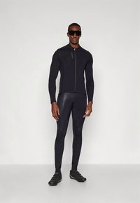 Athlete wearing black full-zip cycling jacket, tight black cycling pants, black cycling shoes, and large black sunglasses standing against plain background.