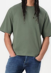 Oliivinvihreä, lyhythihainen puuvillainen t-paita, jossa on väljähkö istuvuus ja pyöreä kaulus, yhdistetty vaaleansinisiin farkkuihin.