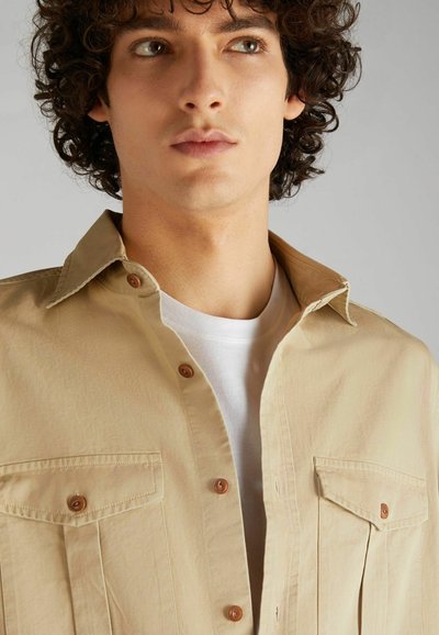 Camicia beige con colletto aperto, due tasche sul petto e bottoni marroni. Indossata sopra una t-shirt bianca a girocollo.