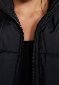 Zwarte gewatteerde jas met een ritssluiting aan de voorkant, met een gladde, glanzende textuur. De kraag is aansluitend en de naden zijn zacht gewatteerd.