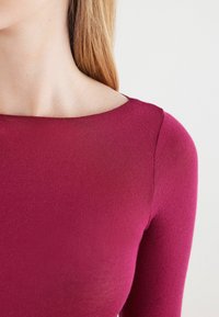 Langærmet top i dyb magenta med en bred udskæring og glat tekstur, der har minimale sømme, hvilket fremhæver den tætsiddende design.
