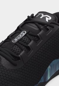 Μαύρο αθλητικό παπούτσι με επάνω μέρος από πλέγμα, μαύρα κορδόνια περασμένα από πλαστικές οπές, ετικέτα "CXT-2" στη γλώσσα και μπλε ιριδίζουσα λεπτομέρεια στη σόλα.