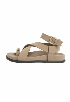 Beige flad sandal med sort sål, med krydsede brede remme, tåloop med guldtone og justerbar ankelrem med metalspænde.