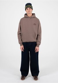 Sudadera con capucha marrón con bolsillo tipo canguro y logotipo, combinada con vaqueros de pierna ancha de color azul oscuro. La modelo lleva un gorro de punto azul oscuro y verde.