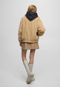 Veste teddy beige et duveteuse avec capuche bleu marine, associée à une jupe beige et des bottes blanches montantes, vue de dos sur un fond neutre.