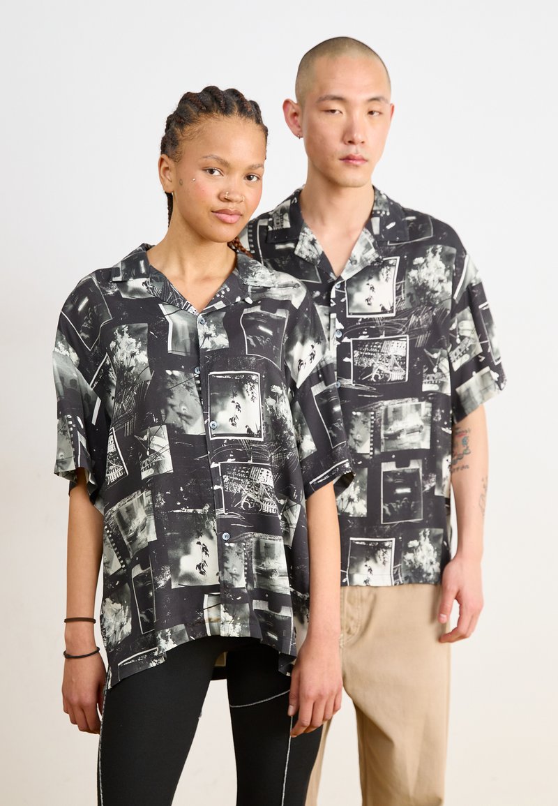 Camicie a manica corta bianco e nero con stampa fotografica. Vestibilità ampia, colletto a scollatura profonda e chiusure con bottoni, indossate con pantaloni casual.