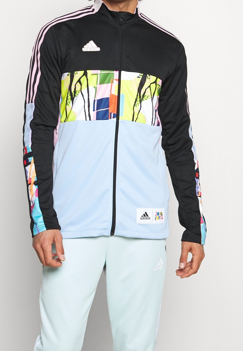 Homme portant une veste zippée Adidas multicolore avec des panneaux noirs, bleu clair et à motifs abstraits, et un pantalon Adidas bleu clair.