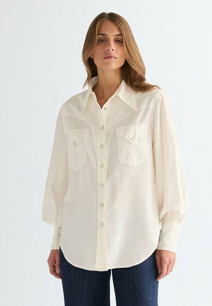 Flødefarvet skjorte med knapper, to brystlommer, klassisk krave og rynkede ærmer; har et blødt, struktureret stof og rundet kant.