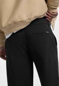 Black pants feature a smooth texture with a slim fit, dual back pockets, and a small logo detail on the right side.