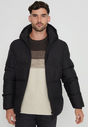 Chaqueta acolchada negra con capucha, con un cierre frontal de cremallera, diseño acolchado y puños elásticos. Lleva puesta sobre un suéter de punto a rayas.