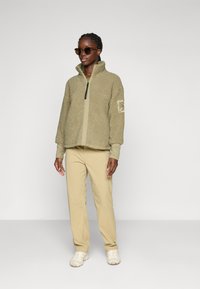 Donna che indossa occhiali da sole, giacca in pile beige, pantaloni beige e sneakers di colore chiaro, in piedi davanti a uno sfondo grigio uniforme.