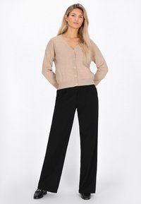 Beige Strickjacke mit V-Ausschnitt, sieben Knöpfen auf der Vorderseite und gerippten Bündchen, kombiniert mit lockeren schwarzen Hosen und schwarzen Schuhen.