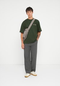 Green cotton t-shirt with embroidered text, grey sling bag, striped grey trousers, and cream sneakers. Simple, relaxed fit design.