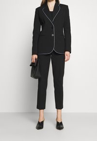 Černý oblek s bílým lemováním, dobře padnoucí sako a úzké kalhoty. Doplněno černou psaníčkovou kabelkou a mules, což podtrhuje elegantní, moderní vzhled.