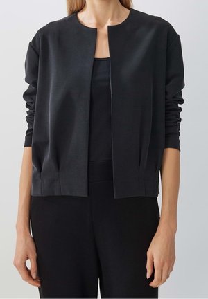 Vrouw met een zwarte, lange mouwen, openvallende cardigan met gerimpelde mouwen, gedragen over een zwart shirt en zwarte broek, tegen een effen achtergrond.