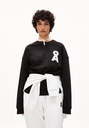 Schwarzer Pullover mit weißem Logo auf der Brust, kombiniert mit weißen Jogginghosen, die ein kleines schwarzes Logo haben. Das Material wirkt weich und lässig.