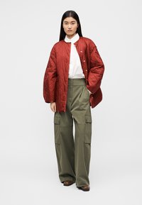 Κόκκινο καπιτονέ μπουφάν με κουμπιά στιγμής, λευκό πουκάμισο και χακί παντελόνι cargo με μεγάλες τσέπες. Καφέ παπούτσια ολοκληρώνουν την εμφάνιση.