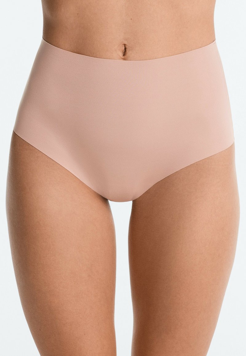 Femme portant des sous-vêtements beige nude taille haute sans coutures, sur un fond clair, montrant le bas du ventre et le haut des cuisses.