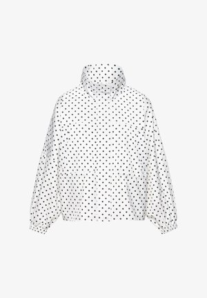 Fehér dzseki magas gallérral és hosszú ujjakkal, egyenletesen elhelyezett kis fekete pöttyökkel borítva, rugalmas mandzsettákkal és elülső zsebekkel.