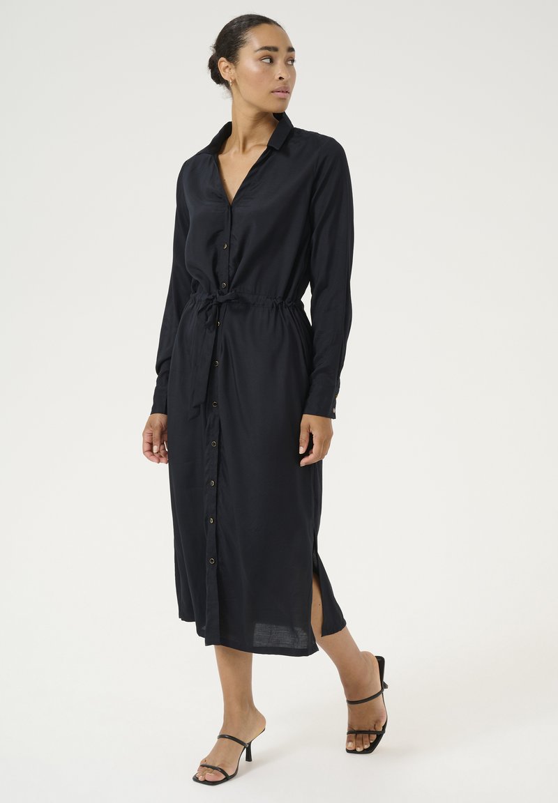 Robe chemise noire à boutons, avec une taille cintrée, des manches longues, des fentes latérales et une texture lisse. Associée à des sandales noires minimalistes.