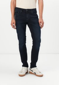 Mörkblå denimjeans med smal passform, utrustade med fem fickor, dragkedja och subtila sömnadsdetaljer. Bärs med vita sneakers.