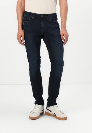 Dark blue denim jeans with a slim fit, five pockets, and a standard button closure, paired with white and black sneakers.