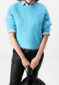 Pull bleu clair en matière pelucheuse avec un col rond et des manches retroussées, porté par-dessus une chemise rayée à col. Associé à un pantalon en cuir noir ajusté.