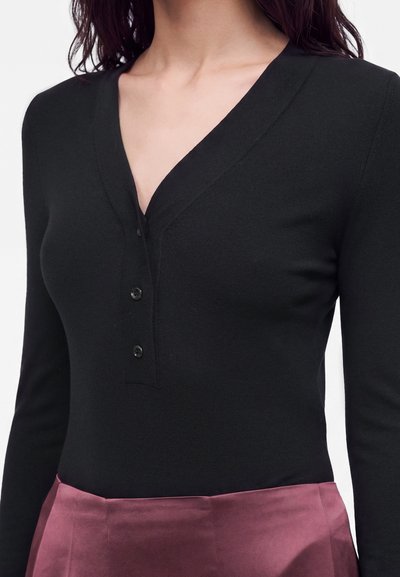 Femme portant un haut noir ajusté à manches longues boutonné avec un décolleté en V et une jupe en satin violette taille haute.