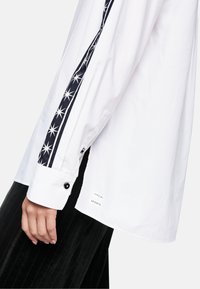 White long-sleeve shirt with black buttoned cuffs and star-patterned black stripe along sleeve, worn with black pleated pants.