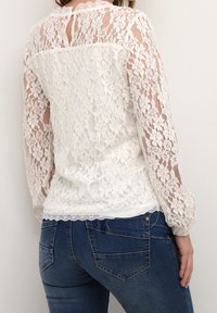 Blusa bianca in pizzo con maniche lunghe, scollatura a onda, apertura a occhiello sul retro, texture stratificata, indossata sopra jeans in denim blu.