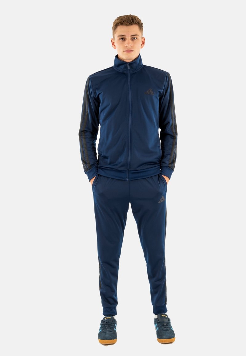 Survêtement bleu marine en tissu lisse avec une veste zippée, des manches longues et des rayures noires contrastantes. Pantalons assortis et chaussures bleues.